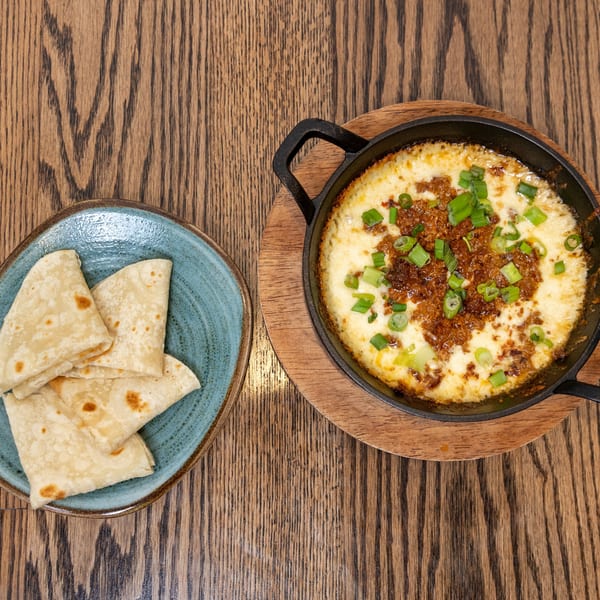 Melted cheese dip with ground meat and green onions in a cast iron pan, with tortillas on a blue plate.