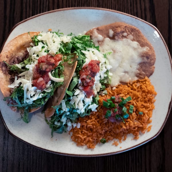 Two La Cocina tacos with lettuce, cheese, salsa, alongside rice and refried beans on a plate.