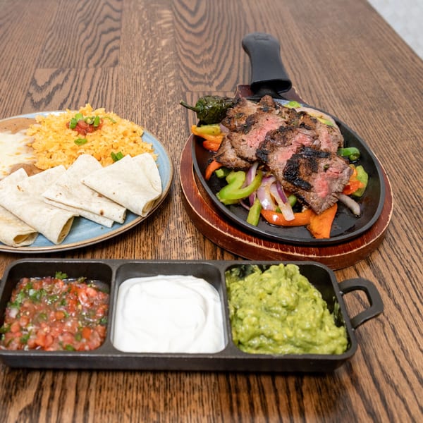 La Cocina's sizzling steak fajitas with rice, tortillas, salsa, sour cream, and guacamole on a wooden table.