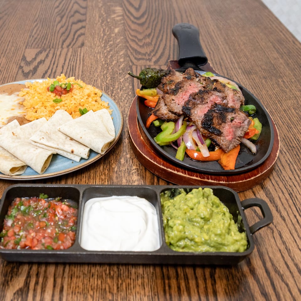 La Cocina's sizzling steak fajitas with rice, tortillas, salsa, sour cream, and guacamole on a wooden table.