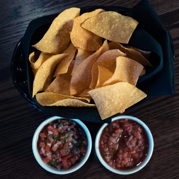 A basket of La Cocina's crispy tortilla chips with two bowls of fresh salsa on a dark wooden table.