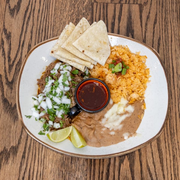 La Cocina's birria plate with shredded beef, rice, beans, tortillas, and lime wedges on a wooden table.